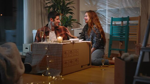 A beautiful couple has their first dinner in their new house, eating pizza and toasting with wine using a box as a table.
He asks her to marry him showing her wedding ring
