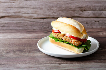 Milanesa sandwich with tomato slices, lettuce and mayonnaise in a white plate on a wooden background with copy space.