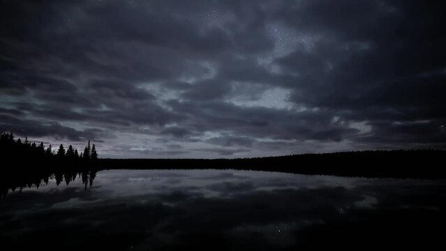 A nighttime time-lapse of the night sky above a calm lake. The clip starts with fast moving clouds that clear to reveal a star filled sky. A few planes streak across the sky.
