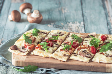 Freshly baked flammkuchen, traditional french tarte flambee or german pizza in a vegetarian recipe with mushrooms, cream cheese, tomatoes and arugula, on light green wooden board and table