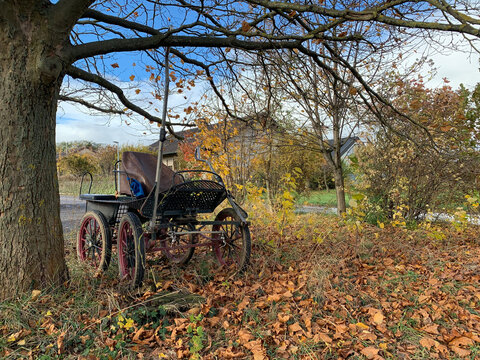 An old open single-seater four-wheeled horse-drawn carriage without a horse. A tarantass or carriage stands under a tree in autumn among fallen leaves in sunny weather in a German village in Germany.