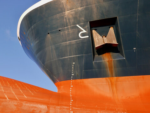 Ship bulbous bow out of water in dry dock. Draft marks on orange red hull. Anchor and rust.