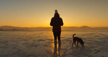 SILHOUETTE: Dog and ice skater in the middle of a frozen lake in golden light