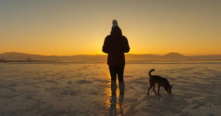 Foto op Aluminium SILHOUETTE: Dog and ice skater in the middle of a frozen lake in golden light © helivideo