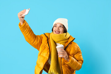 Young Asian woman in winter clothes with coffee cup taking selfie on blue background