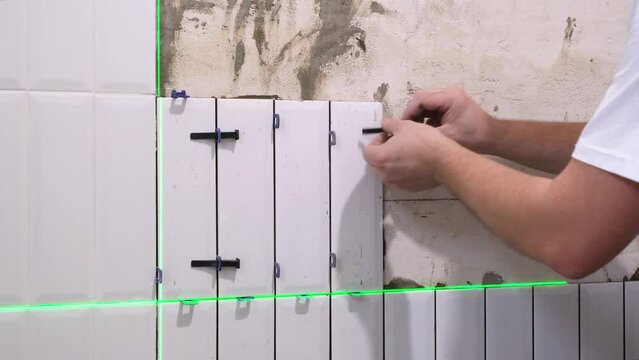 The hands of the tiler are laying white ceramic tile on the wall, using electronic laser level