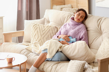 Young woman with hot water bottle sitting on sofa at home - obrazy, fototapety, plakaty