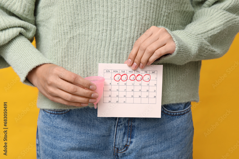 Wall mural Young woman holding menstrual cup and calendar with marked days of menstruation on yellow background