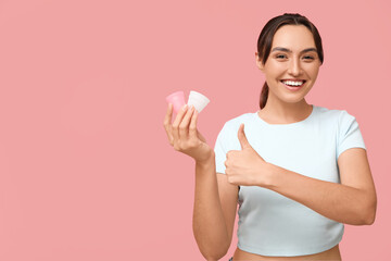 Beautiful young woman with menstrual cups showing thumb-up gesture on pink background