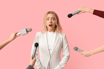 Portrait of stressed young woman surrounded by journalists with microphones on pink background
