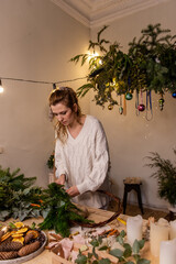 Vertical photo of young woman making handmade Christmas wreath at home from natural materials. DIY...