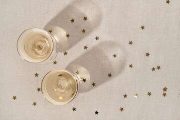 Top view of two wineglasses with sparkling wine on neutral beige linen tablecloth background with natural sunlight shadows and gold star confetti. Festive party event celebration backdrop, copy space