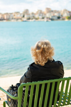 Old Age Alone. Old Woman With Bag Seen From Behind Sitting Looking Out To Sea. Grandmother Loneliness.