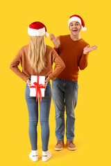 Young woman with Christmas gift and her boyfriend in Santa hats on yellow background