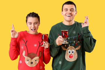 Young couple in Christmas sweaters with mulled wine making wishes on yellow background