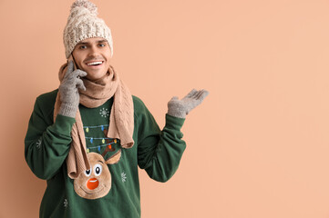 Young man in Christmas sweater talking by mobile phone on beige background