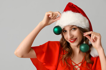 Young woman in Santa hat with Christmas balls on grey background
