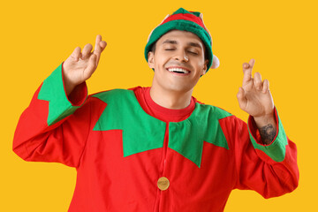 Handsome young man in elf's costume with crossed fingers on yellow background