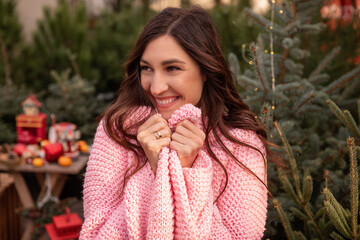 Beautiful young woman in knitted pink sweater, wrapping herself up, brown curly hair blowing in the wind. Smiling girl, close-up portrait against background of green Christmas tree market. Copy space