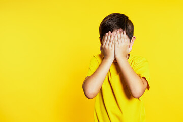 Child in vivid yellow tee stands against a matching backdrop, hands over face in a playful 'see no evil' pose. - obrazy, fototapety, plakaty