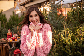 Beautiful young woman in knitted pink sweater, wrapping herself up, brown curly hair blowing in the wind. Smiling girl, close-up portrait against background of green Christmas tree market. Copy space