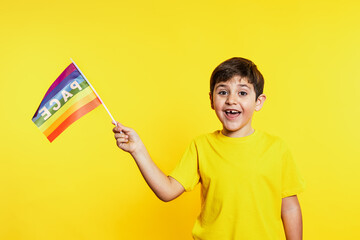 Happy young boy in yellow shirt proudly waves a rainbow peace flag, symbolizing diversity and joy, against a vibrant yellow backdrop.