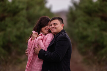 Close-up portrait of happy couple hugging in green coniferous forest. Young man tactilely wrapped black coat around curly-haired woman in pink sweater. Winter engagement, autumn wedding. Marriage