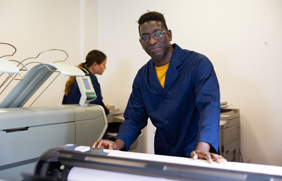 Positive middle-aged African American man in uniform using plotter during work in the printing office