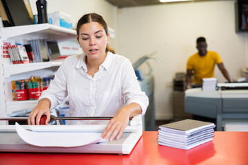 Obraz premium Focused young female typographer trimming the printed work on the cutter in the typography