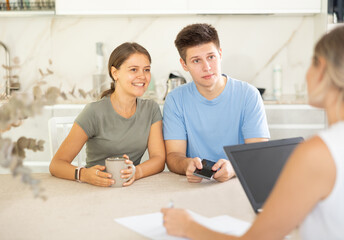 Smiling young couple discussing new home choice with female real estate agent showing options for...