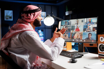 Image showcasing a middle eastern man wearing traditional clothing, speaking with a group of people on his computer screen. An Islamic guy conversing with his team on a conference call. - obrazy, fototapety, plakaty