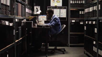 African american private detective walks into archive room with a laptop bag and starts working crime investigation. Male investigator sits at desk with wireless computer, checking suspect case files.