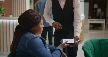 Woman does nfc mobile payment at pos to pay for cup of coffee while waiting on room reservation in hotel lounge area. African american tourist talking to porter about excellent concierge service. - obrazy, fototapety, plakaty