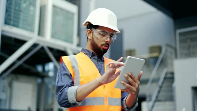 Professional engineer wearing safety helmet and vest standing at the factory and working on a digital tablet. Serious industry manager inspector specialist in uniform looks around checking production