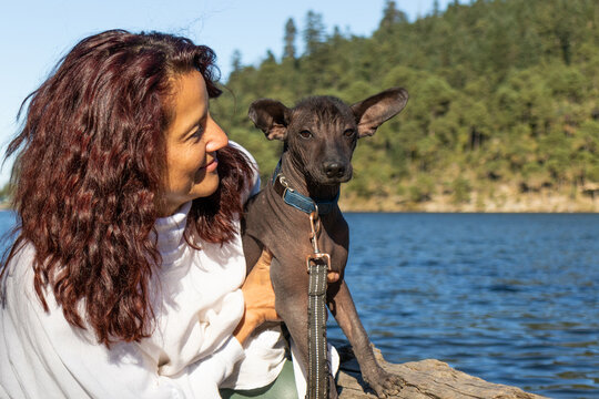 Happy woman sharing a moment of joy with her Xoloitzcuintli dog by a serene blue lake against green forest in daylight
