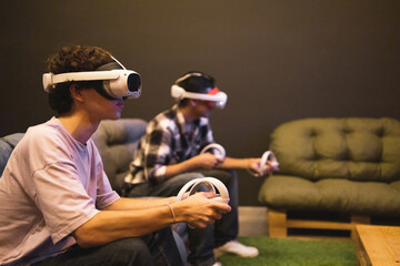 Male friends engrossed in a virtual reality game wearing VR headsets and holding controllers sitting on a couch in a dimly lit room