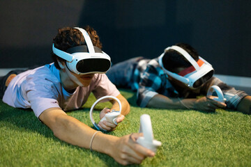 A young man lies prone on artificial grass, engaged in virtual reality gaming with a VR headset and handheld controllers