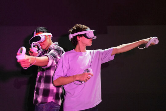 Two friends stand in a gaming area fully immersed in a virtual reality world wearing VR headsets and holding motion controllers in neon light room