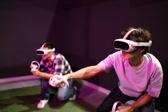 Two friends stand in a gaming area fully immersed in a virtual reality world wearing VR headsets and holding motion controllers in neon light room