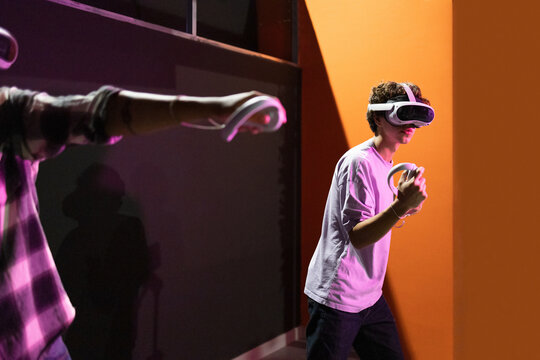 Side view of two friends stand in a gaming area fully immersed in a virtual reality world wearing VR headsets and holding motion controllers in neon light room