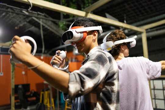 Side view of two friends stand in a gaming area fully immersed in a virtual reality world wearing VR headsets and holding motion controllers