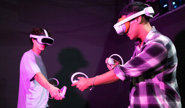 Side view of two friends stand in a gaming area fully immersed in a virtual reality world wearing VR headsets and holding motion controllers in neon light room