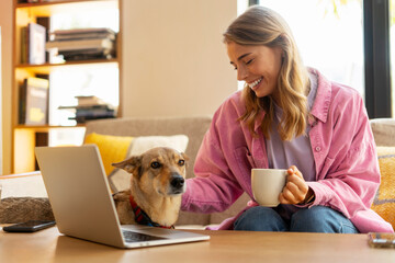 Portrait of smiling attractive woman sitting on sofa using laptop, holding cup, petting dog, at home