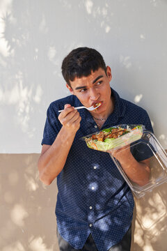 Young man eating Thai salad by green cinder block wall outdoors