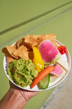 Hand holding plate of chips and guacamole outdoors by green wall