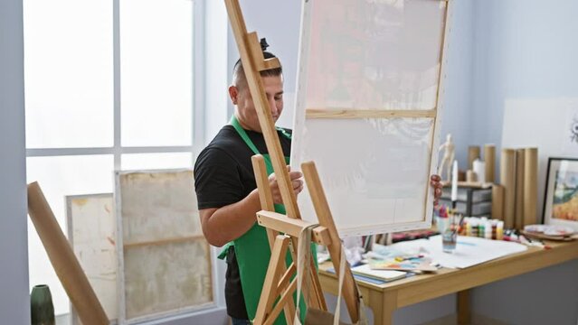 Young, confident latin artist joyfully standing by his easel in the art studio, ready to transfer creativity onto canvas