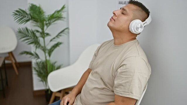 Young latin man, with a serious expression, relaxed and thoroughly engrossed in the audio of his music, sitting in a waiting room, enjoying his own world indoors.
