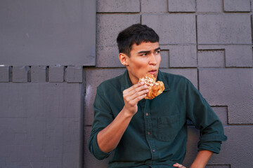 Young man eating flaky pastry by gray geometric brick wall