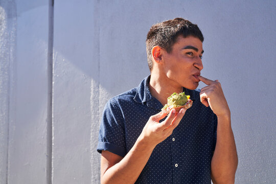 Young man eating avocado toast outdoors by gray textured wall 