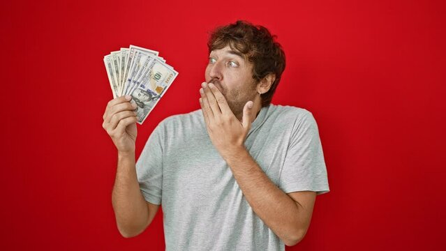 Young blond bearded guy, shocked and quiet, covering mouth with hand, surprised by a secret regarding american us banknotes. fear and shame expressed over isolated red background.